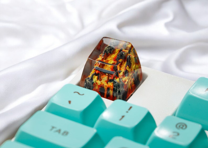 K&ouml;nnte beinhalten: Nahaufnahme eines Tastaturkappen mit einem Harzdesign, das ein feuriges orange-schwarzes Muster zeigt. Die Tastaturkappe befindet sich auf einer wei&szlig;en Tastatur mit blauen Tasten.