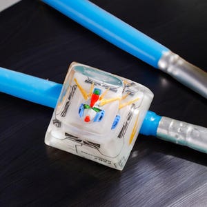 May include: A clear resin cube with a detailed model of a robot inside. The cube is sitting on a black surface with two blue and silver paintbrushes.