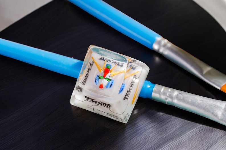May include: A clear resin cube with a detailed model of a robot inside. The cube is sitting on a black surface with two blue and silver paintbrushes.