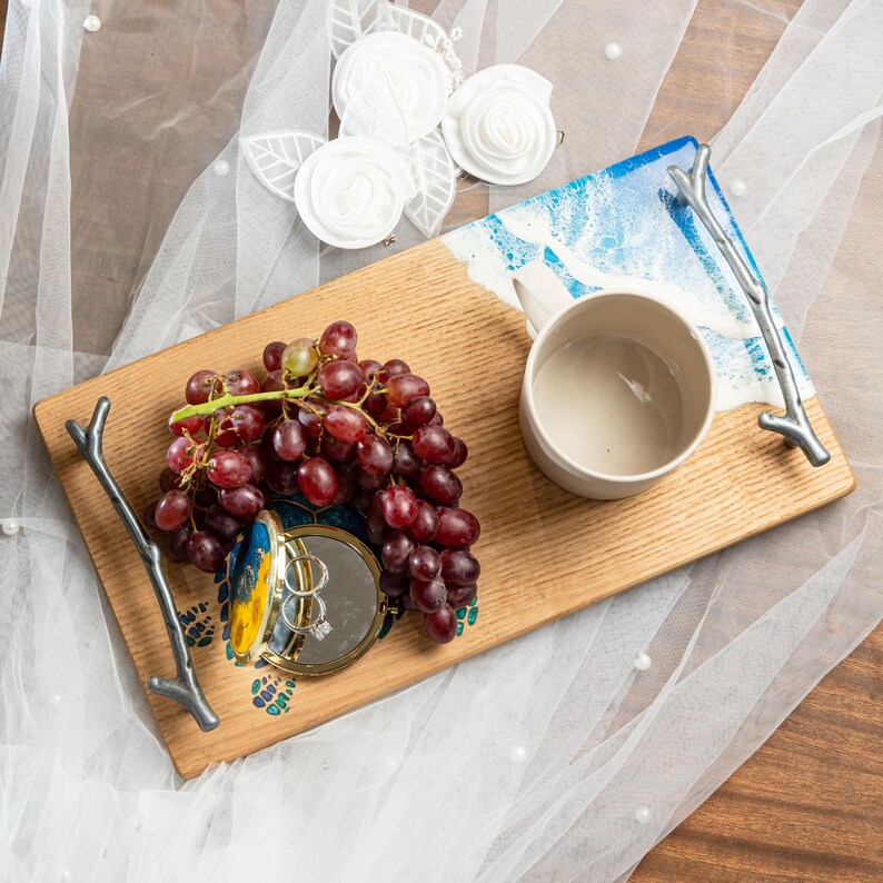 May include: A wooden serving tray with a blue and white resin inlay. The tray has two twig handles and features a white ceramic mug, a bunch of red grapes, and a small, open, round, gold-colored box with two rings inside.