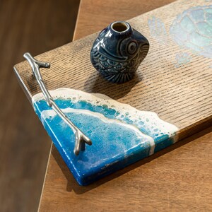 May include: A wooden serving tray with a blue and white resin ocean wave design. The tray has a silver metal branch handle and a blue and white ceramic fish figurine.