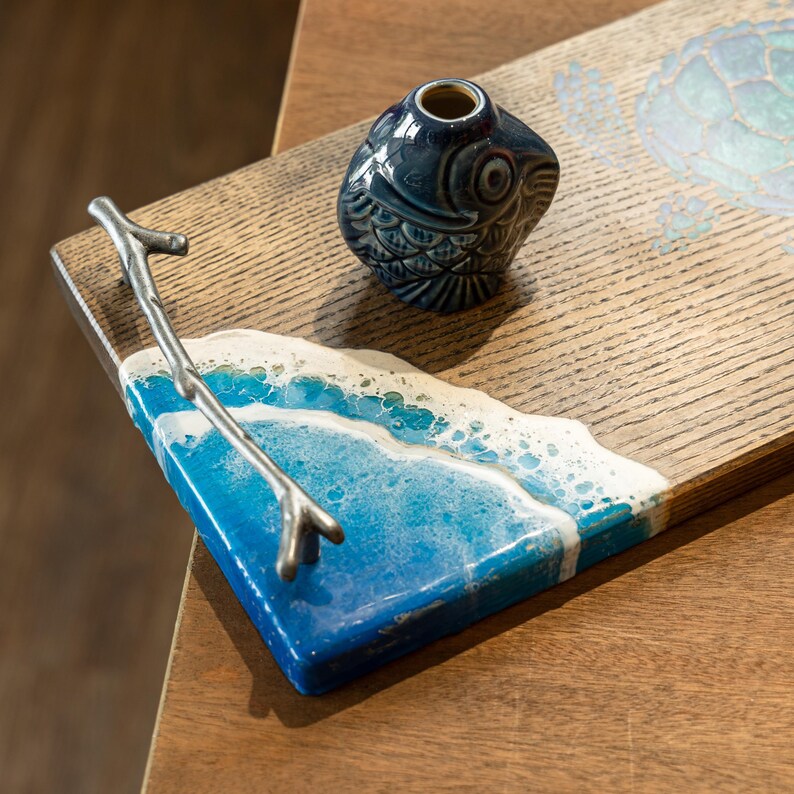 May include: A wooden serving tray with a blue and white resin ocean wave design. The tray has a silver metal branch handle and a blue and white ceramic fish figurine.