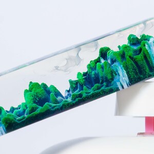 May include: A clear glass tube filled with a green and blue liquid that resembles a landscape. The liquid is moving and swirling inside the tube.