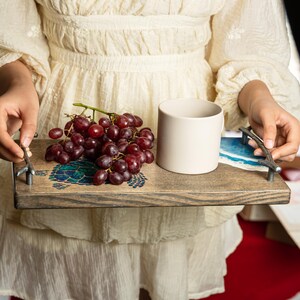 May include: A wooden serving tray with metal handles features a blue and green turtle design. A white ceramic mug and a bunch of red grapes are on the tray.