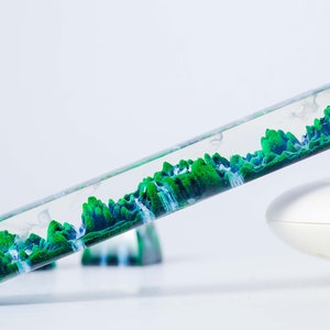 May include: A clear glass tube filled with green and blue liquid, resembling a landscape with mountains and waterfalls. The tube is tilted at an angle, and the liquid is flowing.