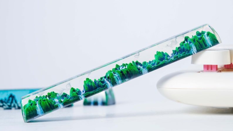 May include: A clear glass tube filled with green and blue liquid, resembling a landscape with mountains and waterfalls. The tube is tilted at an angle, and the liquid is flowing.