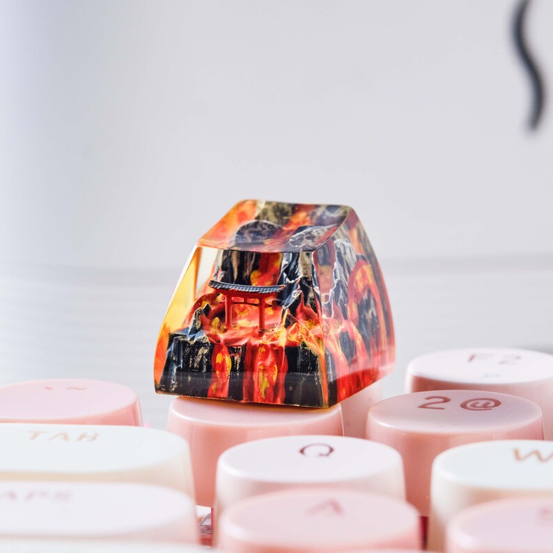 K&ouml;nnte beinhalten: Eine rote, orange und schwarze Harz-Tastenkappe mit einem Miniatur-japanischen Tempel und einer Bergszene. Die Tastenkappe liegt auf einer rosafarbenen Tastaturtaste.