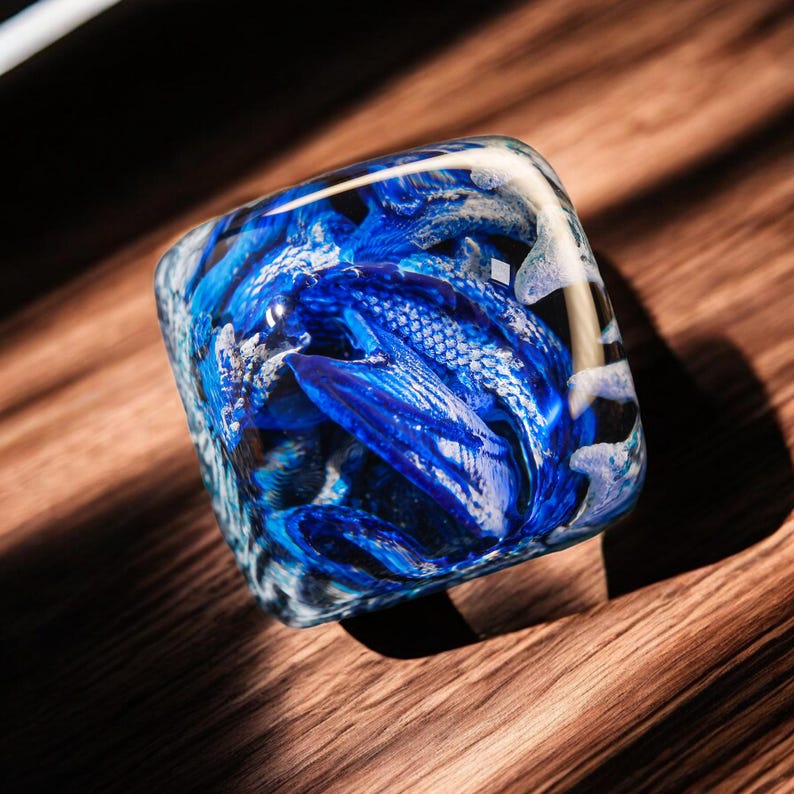 May include: A blue and white glass paperweight with a swirling, abstract design. The paperweight is shaped like a cube and has a smooth, polished surface.