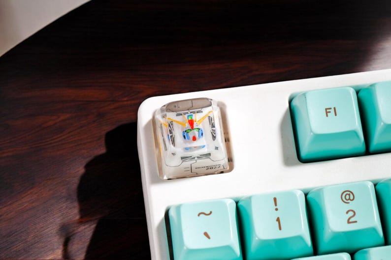 May include: A white keyboard with teal keys. The top left key has a clear keycap with a colorful design of a race car. The keys are labeled with symbols such as "~", "!", "@", and "F1".