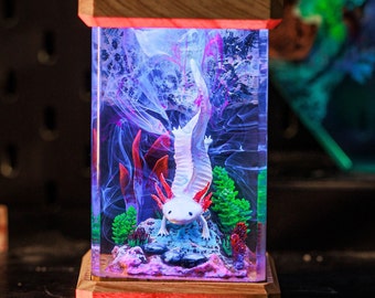 May include: A white axolotl figurine with pink gills is encased in a clear resin lamp base. The lamp base is made of wood and has a red light inside. The axolotl is surrounded by blue and purple resin swirls and green and red plants.