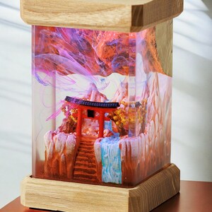 Op de afbeelding: Een houten doos met een transparante harsinzet met een miniatuur Japans tafereel. De sc&egrave;ne bevat een rode torii-poort, een waterval en bergen. De hars is gevuld met roze en blauwe swirls.