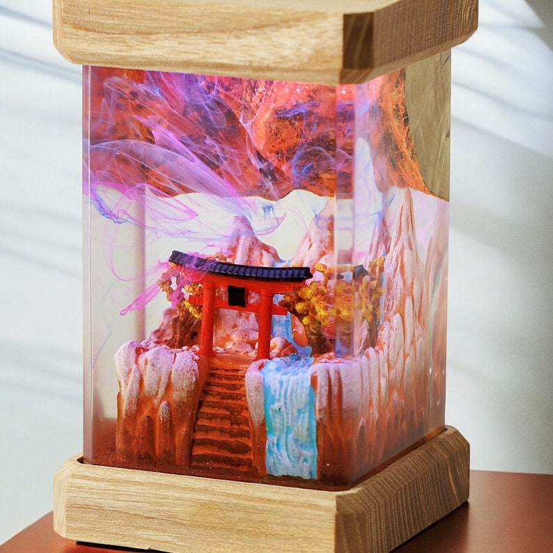 Op de afbeelding: Een houten doos met een transparante harsinzet met een miniatuur Japans tafereel. De sc&egrave;ne bevat een rode torii-poort, een waterval en bergen. De hars is gevuld met roze en blauwe swirls.