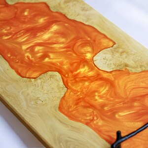 May include: A wooden serving tray with a large, orange resin inlay. The resin has a swirling, marbled pattern and a glossy finish. The tray has a black metal handle.