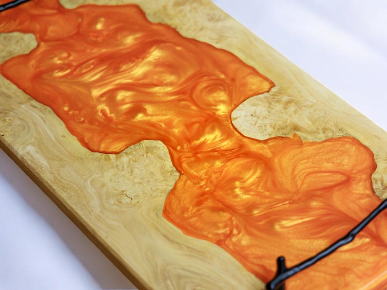May include: A wooden serving tray with a large, orange resin inlay. The resin has a swirling, marbled pattern and a glossy finish. The tray has a black metal handle.