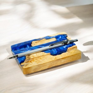 May include: A wooden pen holder with a blue resin inlay. The holder has a groove for a pen and is made of natural wood with a light brown color.