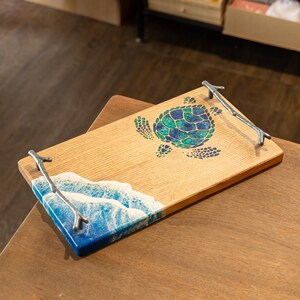 May include: A wooden serving tray with a blue and green resin wave and turtle design. The tray has two silver metal twig handles.