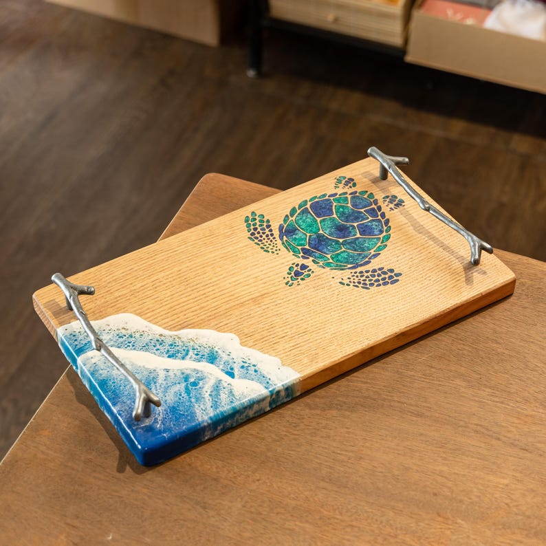 May include: A wooden serving tray with a blue and green resin wave and turtle design. The tray has two silver metal twig handles.