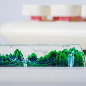 May include: A clear glass rectangular prism filled with green and blue sand, creating a landscape scene. The sand is arranged in a mountainous formation with a waterfall flowing down the side.