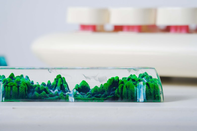 May include: A clear glass rectangular prism filled with green and blue sand, creating a landscape scene. The sand is arranged in a mountainous formation with a waterfall flowing down the side.