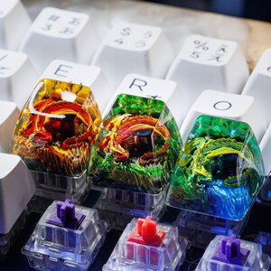May include: Three custom keyboard keycaps with dragon designs encased in clear resin. The keycaps are yellow, green, and blue with red and gold accents. The keycaps are on a white keyboard.