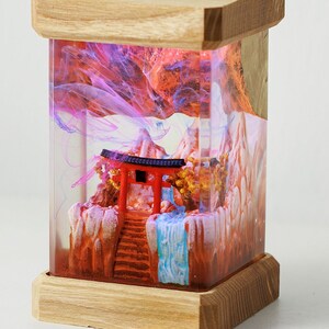 Op de afbeelding: Een houten doos met een glazen voorzijde die een miniatuur sc&egrave;ne bevat van een rode torii-poort, een waterval en bergen. De sc&egrave;ne wordt van achteren verlicht met roze en blauw licht.
