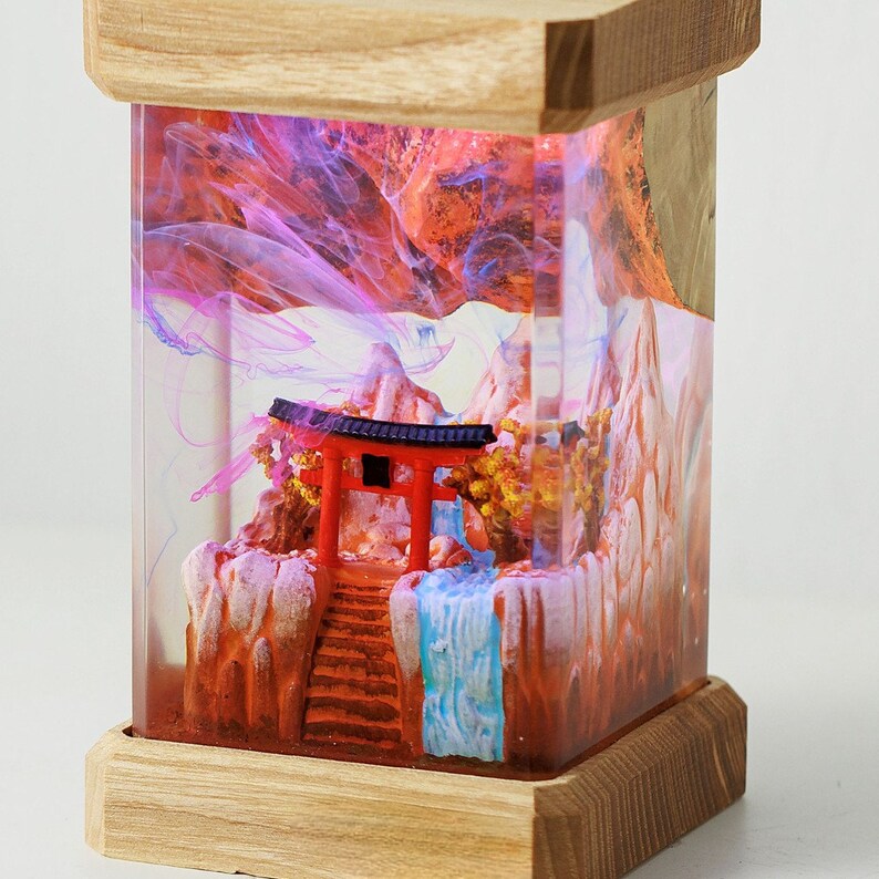 Op de afbeelding: Een houten doos met een glazen voorzijde die een miniatuur sc&egrave;ne bevat van een rode torii-poort, een waterval en bergen. De sc&egrave;ne wordt van achteren verlicht met roze en blauw licht.