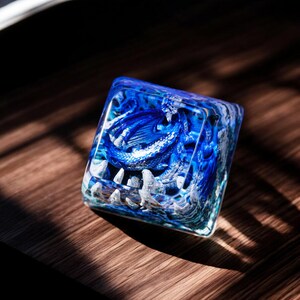 May include: A blue and white resin keycap with a detailed design of a dragon inside. The keycap is clear and shows the dragon in detail.