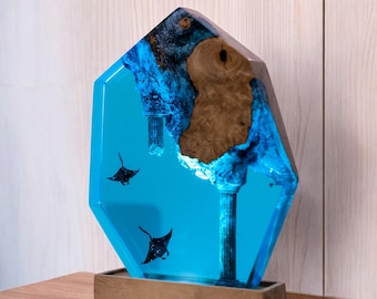 May include: A decorative resin sculpture featuring a geometric shape with a light blue interior. Two black manta ray silhouettes swim within the blue resin. A section of brown wood is embedded in the resin, and the sculpture sits on a rectangular base.