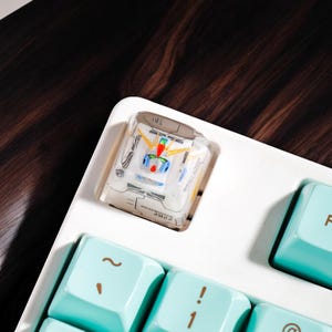 May include: A white keyboard with a teal keycap that says "F1" and a clear keycap with a colorful design. The keyboard is on a dark wood surface.