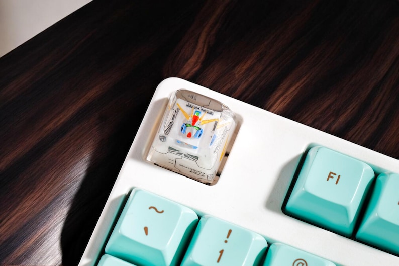 May include: A white keyboard with a teal keycap that says "F1" and a clear keycap with a colorful design. The keyboard is on a dark wood surface.