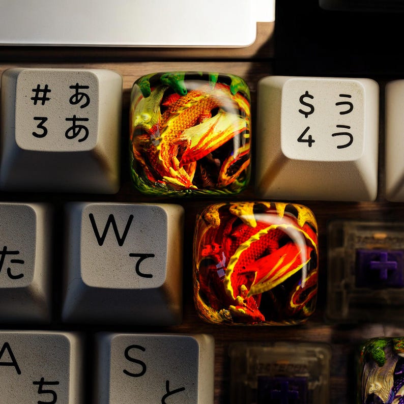 May include: A keyboard with custom keycaps featuring a dragon design. The keycaps are made of resin and have a clear, glossy finish. The dragon design is in shades of red, orange, and yellow. The keycaps are labeled with the letters "W", "S", "A", "#", "$", "3", and "4". The keycaps also have Japanese characters on them.