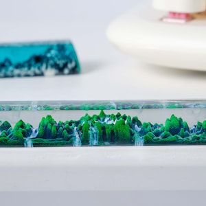 May include: A clear resin keyboard wrist rest with a green and blue landscape design. The design features a mountain range with waterfalls.