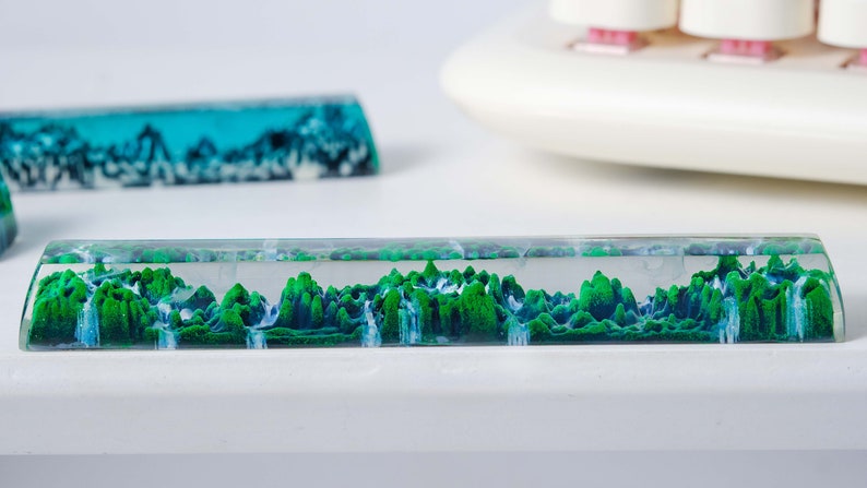 May include: A clear resin keyboard wrist rest with a green and blue landscape design. The design features a mountain range with waterfalls.
