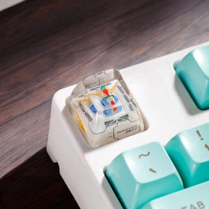 May include: A white mechanical keyboard with a teal keycap set. The keyboard has a custom keycap with a clear plastic design and a printed circuit board inside. The keycap is labeled "2022".
