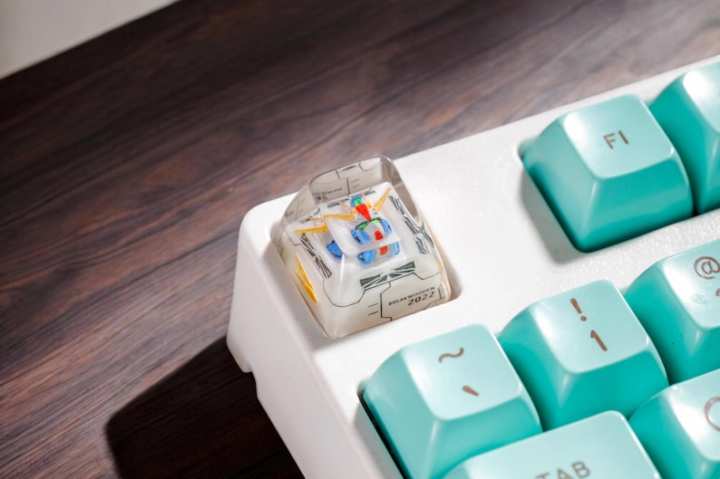May include: A white mechanical keyboard with a teal keycap set. The keyboard has a custom keycap with a clear plastic design and a printed circuit board inside. The keycap is labeled "2022".