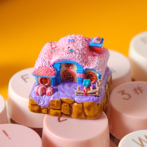 May include: A miniature pink and white house with blue accents sits on a keyboard. The house has a pink roof with white stars and a blue chimney. The house is surrounded by a purple base with a brown rock base.
