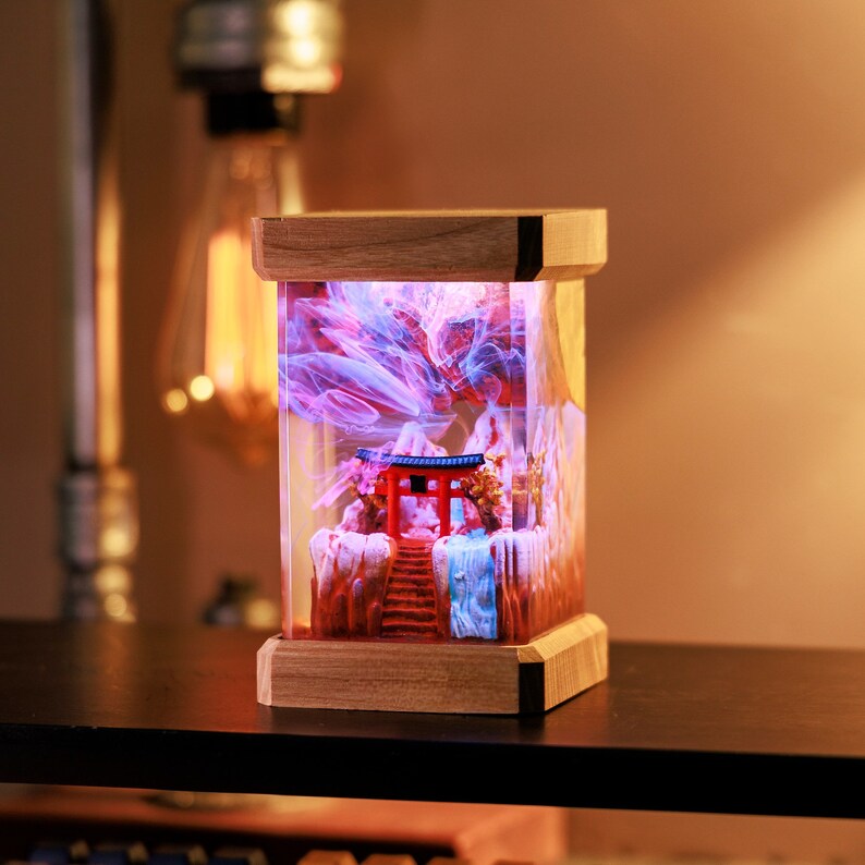 Op de afbeelding: Een houten lampenvoet met een transparante glazen cilinder die een miniatuur rode Japanse tempel en een wervelende paarse en blauwe hars bevat. De hars lijkt op rook.