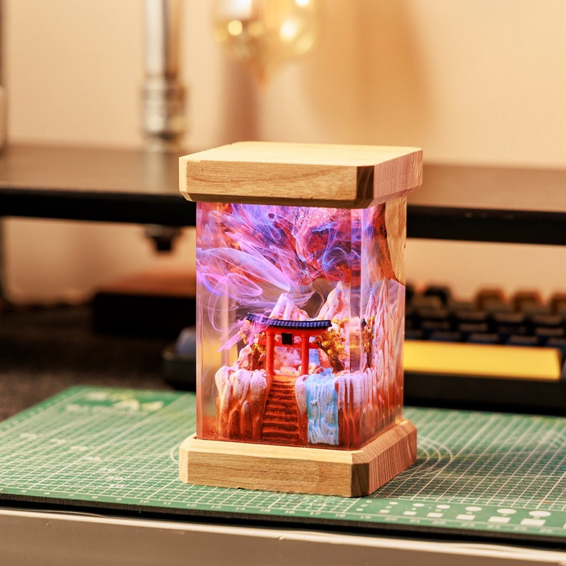Op de afbeelding: Een houten lampenvoet met een transparante harsinzet met een miniatuur Japanse tempel, bergen en wervelende roze en blauwe rook. De lamp wordt van binnenuit verlicht en geeft een warm gloed.