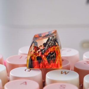 K&ouml;nnte beinhalten: Eine transparente Harztastenkappe mit einem roten, orangefarbenen und schwarzen Landschaftsdesign. Die Tastenkappe liegt auf einer rosa Tastatur.