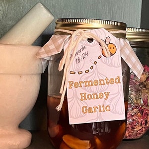 May include: A glass jar with a gold lid and a white and brown checkered cloth tied around the top. The jar contains a fermented honey garlic mixture. A tag attached to the jar reads "Fermented Honey Garlic" and "Proverbs 16:24".