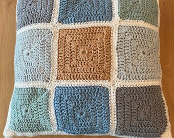 May include: A square crocheted pillow with a patchwork design. The pillow is made of nine squares in different colors, including light blue, gray, beige, and green.