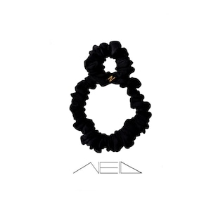 Könnte beinhalten: Schwarzes Samt-Haargummi mit einem goldfarbenen "N"-Anhänger. Das Haargummi hat die Form einer Acht. Das Wort "NEED" steht unter dem Haargummi in einer minimalistischen Schriftart.