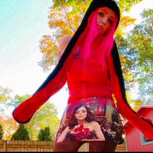 Pu&ograve; includere: Una persona indossa una maglia rossa e una sciarpa con cappuccio nero con una gonna a stampa grafica che presenta una donna con un vestito rosso. La gonna ha la scritta "Never Give Up" stampata.