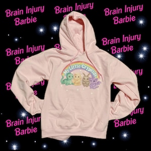May include: A light pink hooded sweatshirt with a front pocket and drawstring. The front features a rainbow with the text "My Little Cryptids" and four cartoon creatures. The words "Brain Injury Barbie" are repeated around the image.