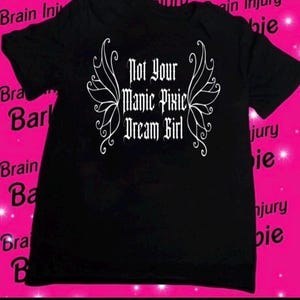 Puede incluir: Camiseta negra con un diseño gráfico blanco de un par de alas y el texto "Not Your Manic Pixie Dream Girl".