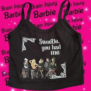 Może przedstawiać: Czarny top na ramiączkach z napisem "Sweetie, you had me" i grafiką kilku postaci. Tło jest różowe ze słowami "Brain Injury Barbie" powtórzonymi. Top ma cienkie ramiączka.