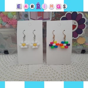 May include: Two pairs of earrings are displayed on white cards. One pair features white daisy-shaped beads with yellow centers. The other pair has a colorful, rectangular design with rainbow-colored beads. The word "EARRINGS" is spelled out above the earrings.