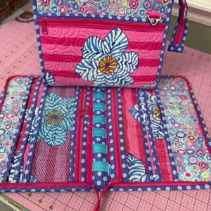 May include: A quilted fabric organizer set featuring a rectangular bag and a matching fold-out wallet. Both items display a vibrant pattern of pink stripes, blue polka dots, and a multi-colored floral print with a large blue and white flower applique.
