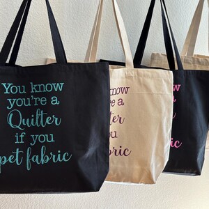 May include: Three tote bags with different colored fabric. The black bag has the text "You know you're a Quilter if you pet fabric" in white and teal lettering. The beige bag has the text "now you're a quilter if you pet fabric" in purple lettering. The black bag has the text "now you're a quilter if you pet fabric" in pink lettering.