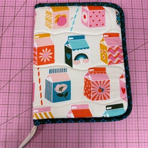 Könnte beinhalten: Eine rechteckige Stofftasche mit Reißverschluss und einem wiederkehrenden Muster aus bunten Milchkartons auf cremefarbenem Hintergrund. Die Kartons zeigen verschiedene Designs wie Blumen, Herzen, Regenbögen und geometrische Formen.